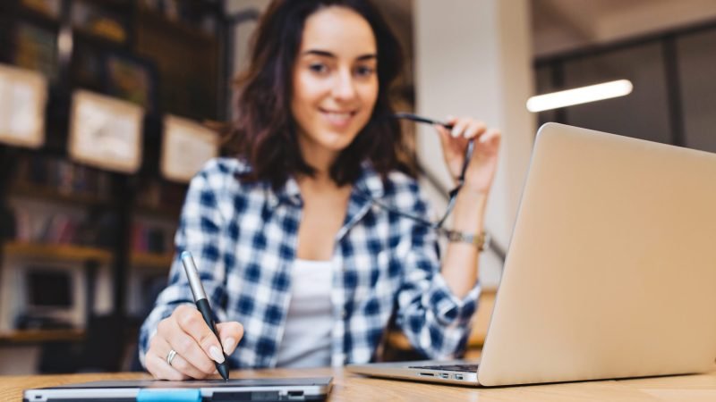 Modern stylish image of smart brunette young woman working with laptop on table in library. Smiling to camera, playing with black glasses, great success, hard-working student. Revalidación de materias