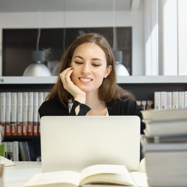 Undergraduate student girl preparing for exams, working on laptop, using wireless Internet connection while sitting at desk with huge stacks of books at college library, resting her elbow on table Revalidación de materias
